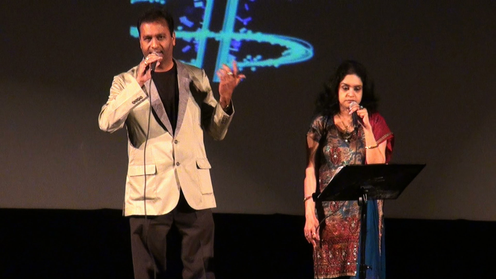A man gesturing while speaking on stage next to a woman