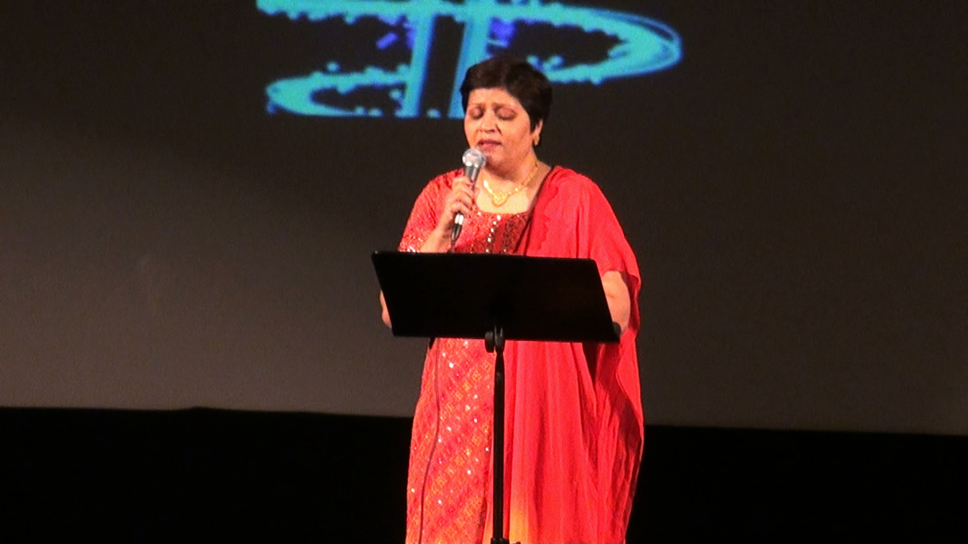 A woman in a red sari singing, reading from sheet music