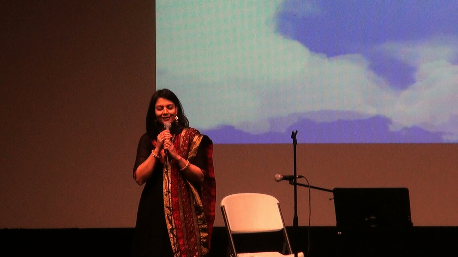 A woman singing with hands folded, in front of a projected screen