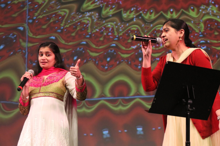 Two women singing a duet in front of a colorful backdrop