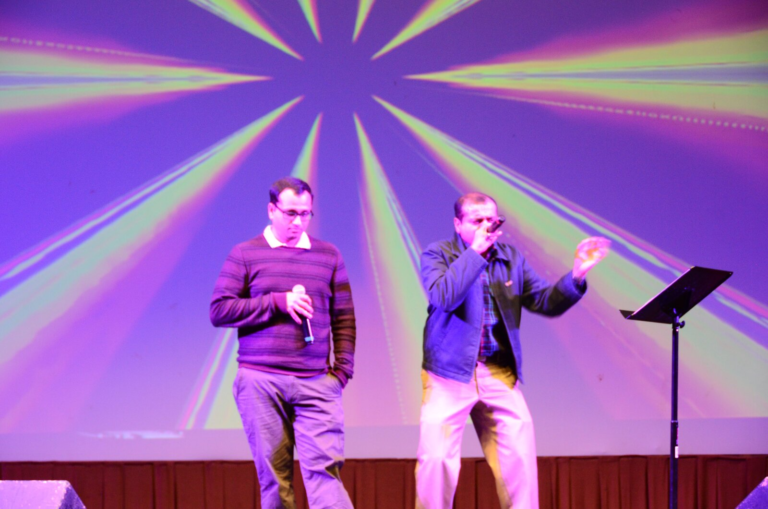 Two men singing together in front of a colorful sunburst backdrop