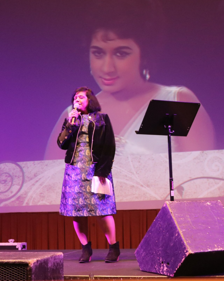A woman singing with a black-and-white film scene projected behind her