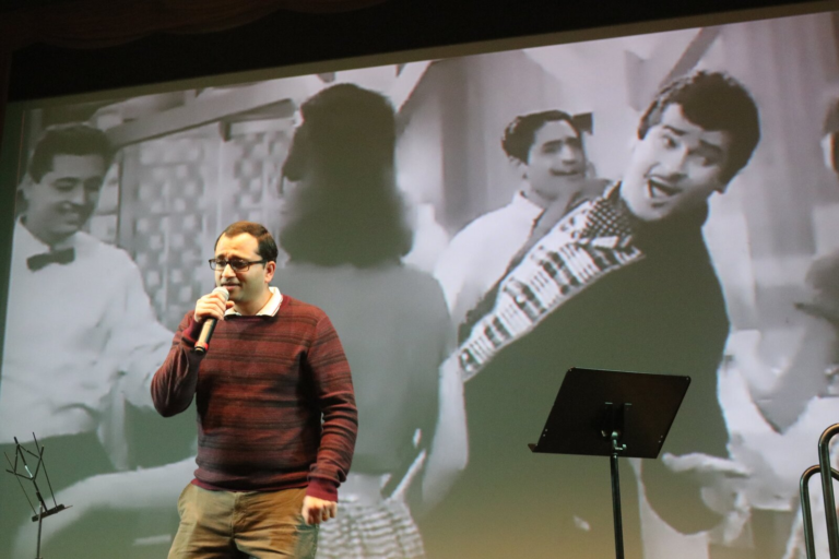 A man singing with a classic film scene projected behind him