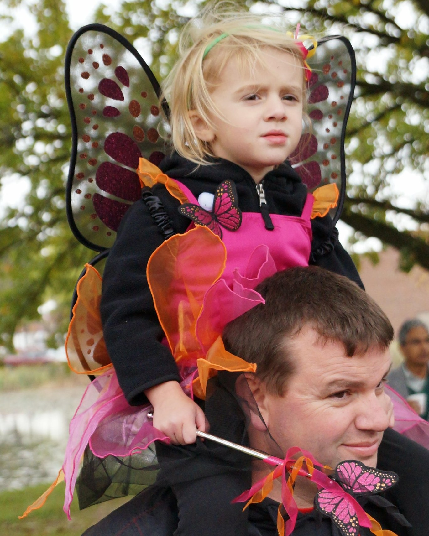A girl in a monarch butterfly costume, holding her butterfly wand, riding on her father's shoulders at the playground dedication