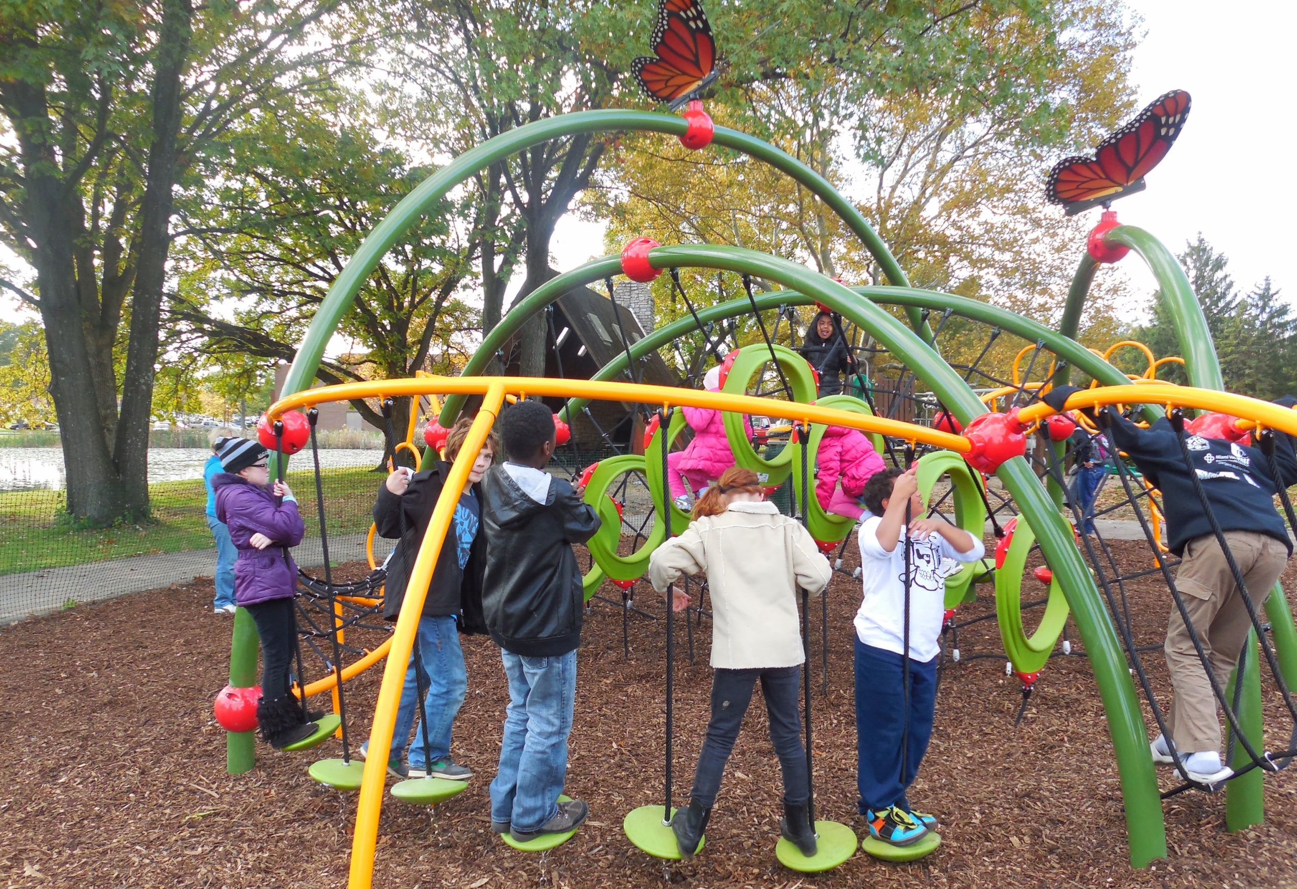 A diverse group of children climbing the arched play structure at the Butterfly Meadow dedication, with two monarch butterfly sculptures overhead
