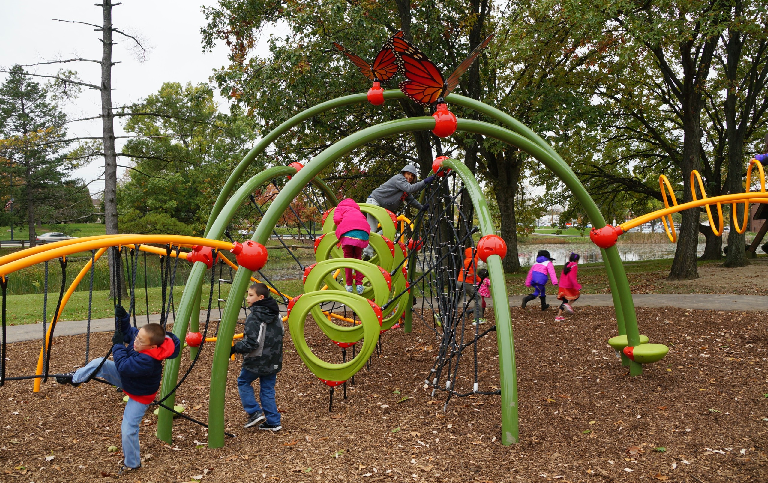 A diverse group of children playing across the arched climbing structure at the Butterfly Meadow dedication, with monarch butterfly sculptures on top and the park pond behind them
